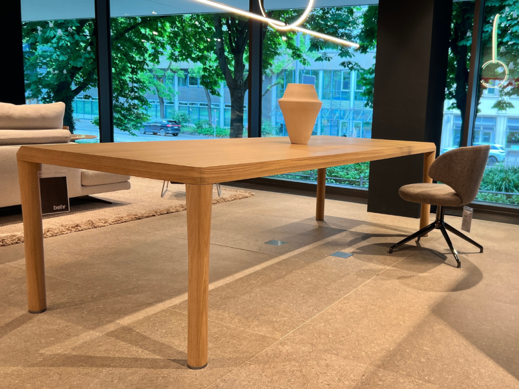 Oak rectangular dining table in a showroom with a beige vase, a gray swivel chair, and a sofa beside large windows.