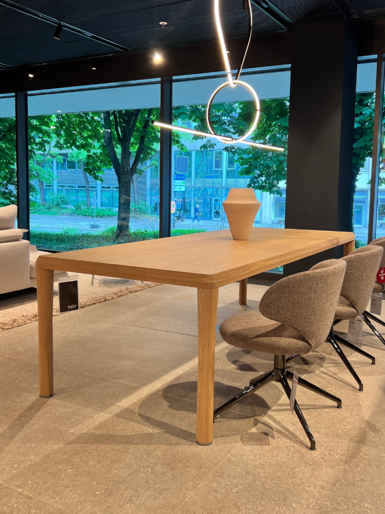 Wood dining table with four legs and beige upholstered chairs, a ceramic vase on the table, in a showroom with large windows overlooking trees.