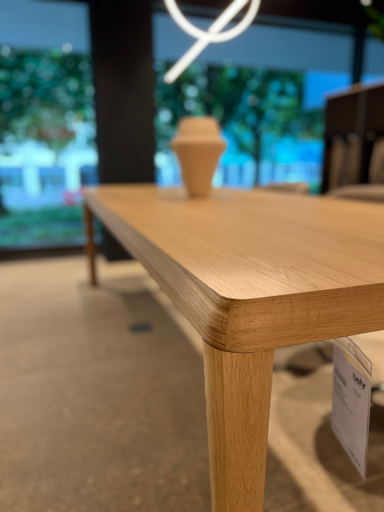 Close-up of a light oak wooden table edge in a showroom, with a blurred beige decorative item and a price tag in the background.
