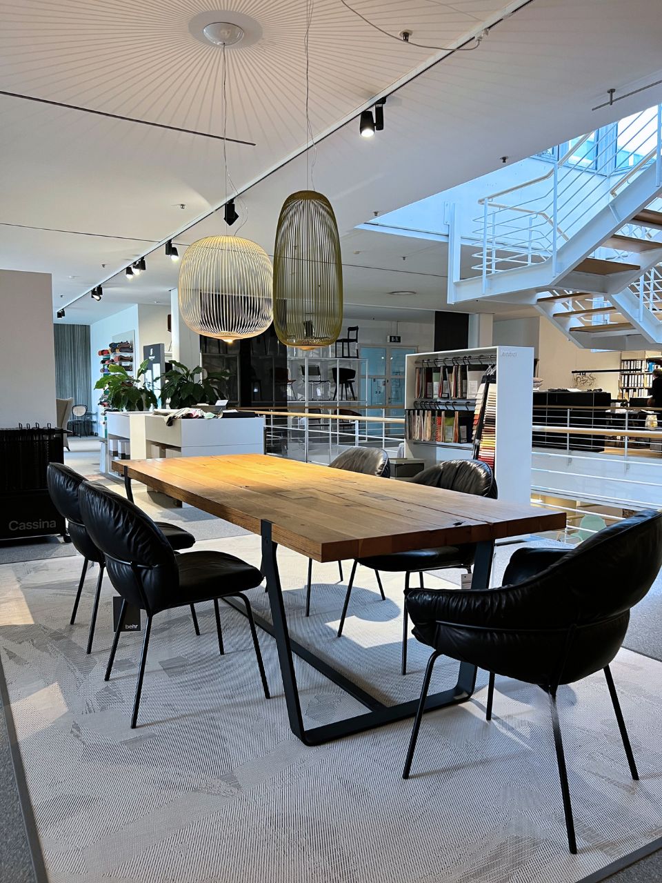 Open-concept furniture showroom with a large wooden dining table surrounded by black leather chairs and two large pendant lamps overhead.
