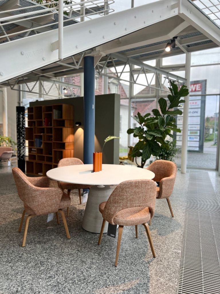 Round white table with an orange vase and a single leaf, surrounded by pink upholstered chairs in a modern showroom setting.