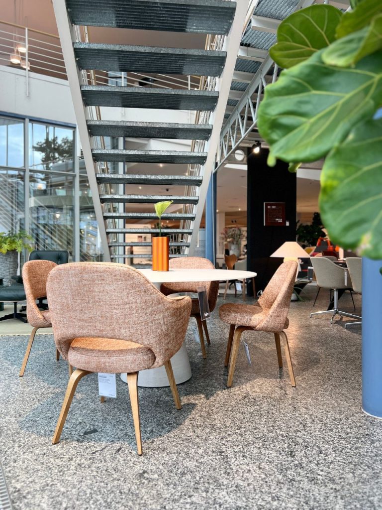 Modern lounge area with a round white table, blush upholstered chairs, and an orange vase with a small plant under a metal staircase.