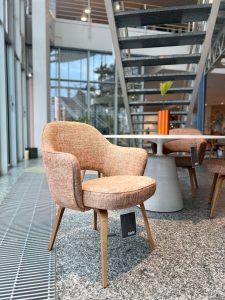 Powder-pink upholstered lounge chair with curved arms and wooden legs beside a round white table in a bright showroom.