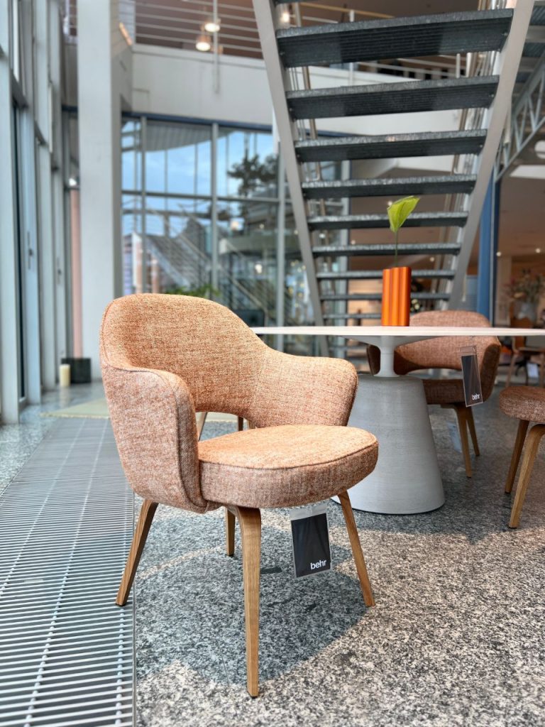 Powder-pink upholstered lounge chair with curved arms and wooden legs beside a round white table in a bright showroom.