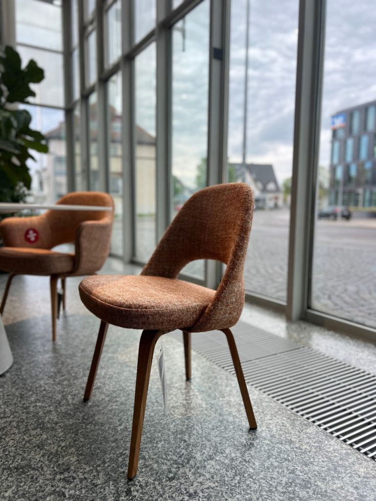 Brown textured upholstered chair with curved back and wooden legs in a bright showroom by large glass windows, with a second chair in the background.