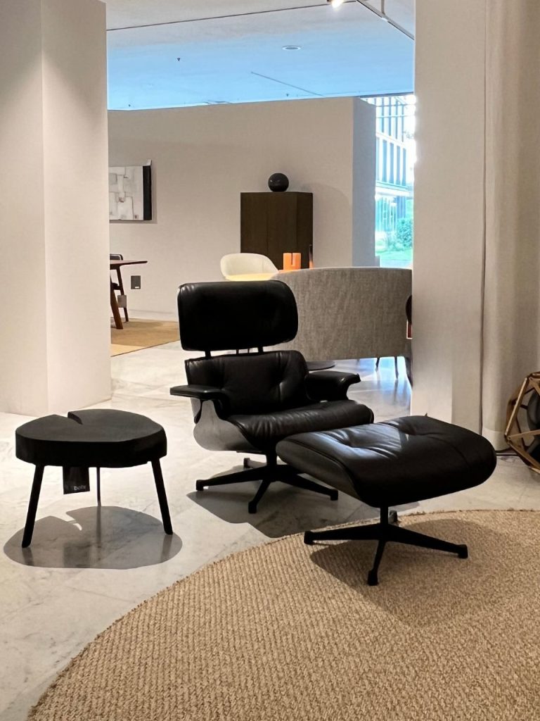 Black leather Eames-style lounge chair with matching ottoman in a modern showroom, next to a small round black table on a marble floor and beige rug.
