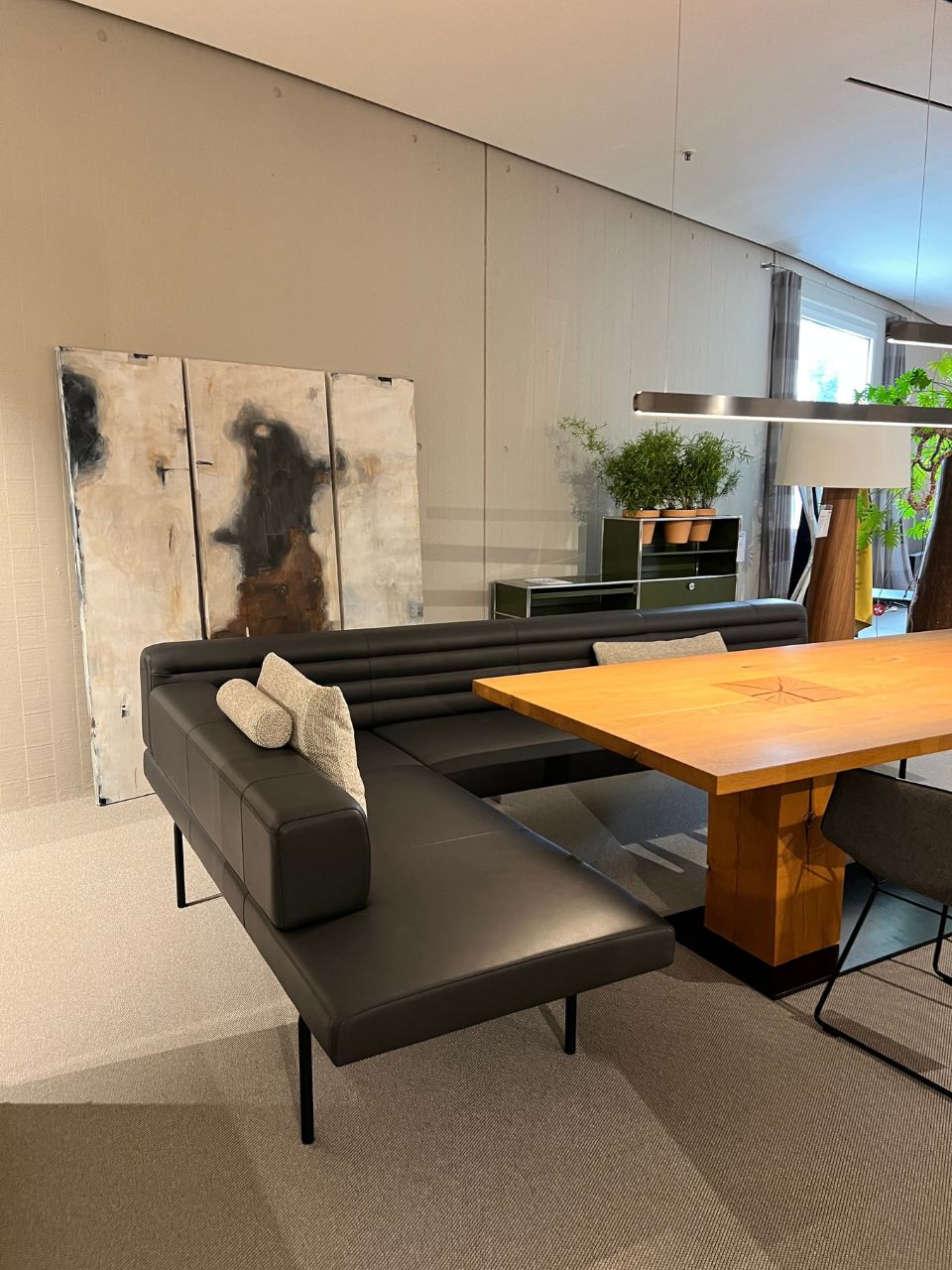 Modern living space with a black L-shaped leather sofa around a large wooden table and abstract artwork propped against a light wall.