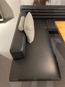 Black leather sectional sofa with two light gray textured cushions in a modern lounge area near a wooden table.