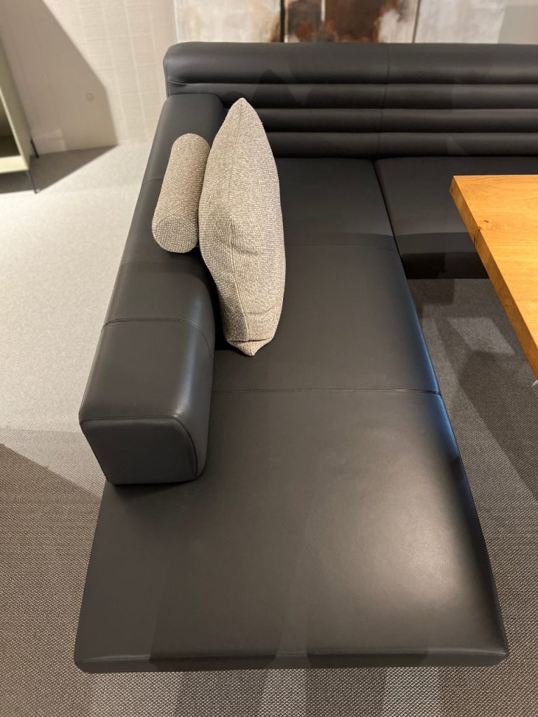 Black leather sectional sofa with two light gray textured cushions in a modern lounge area near a wooden table.