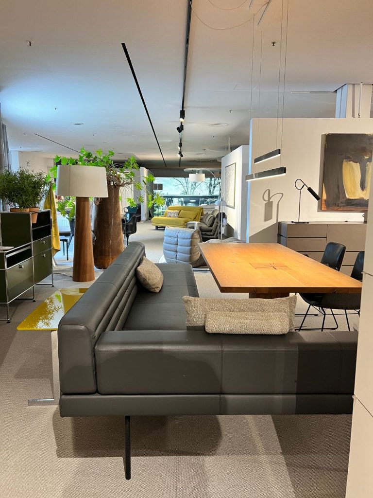 Modern showroom with a large dark gray leather sectional and a rectangular wooden dining table, surrounded by plants and other furniture pieces in the background.