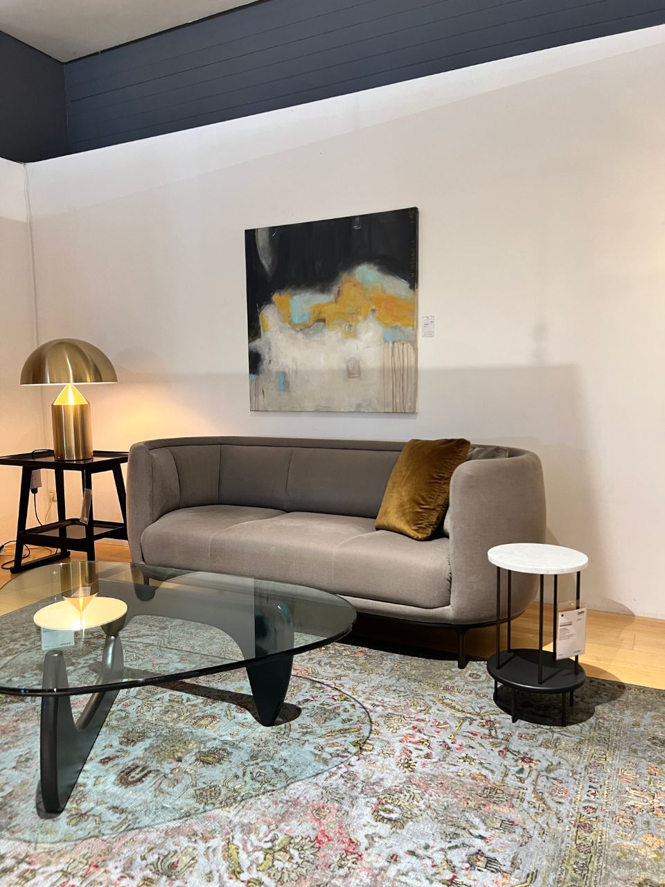 Modern living room display with a gray sofa, mustard throw pillow, glass oval coffee table, and a gold lamp on a side table beside the sofa; abstract painting on the white wall and a patterned area rug.