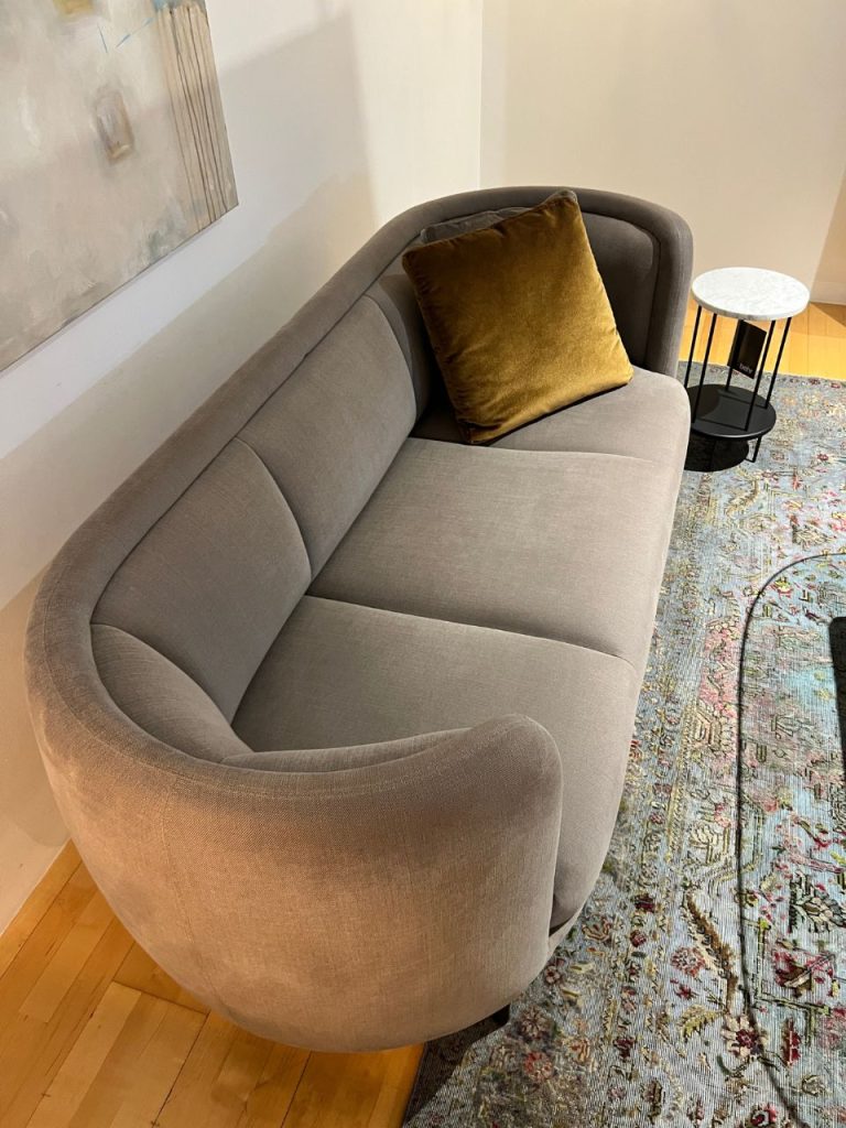 Gray curved sofa with a mustard-yellow throw pillow, beige wall, and a round marble-top side table in a living room.