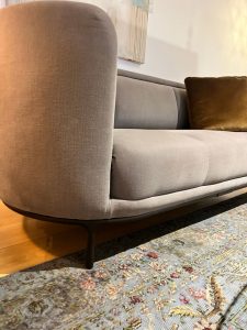 Close-up of a light gray curved-arm sofa with a brown throw pillow, black legs, on a patterned area rug.