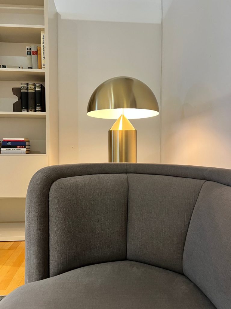 Grey curved sofa in a bright living room, with a gold dome lamp on a side table and a white bookshelf in the background and books on the shelves.