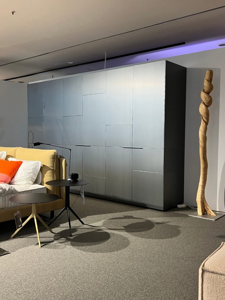 Modern showroom scene with a grey modular storage wall, beige sofa, and black round nesting tables beside a tall, twisted wooden sculpture.