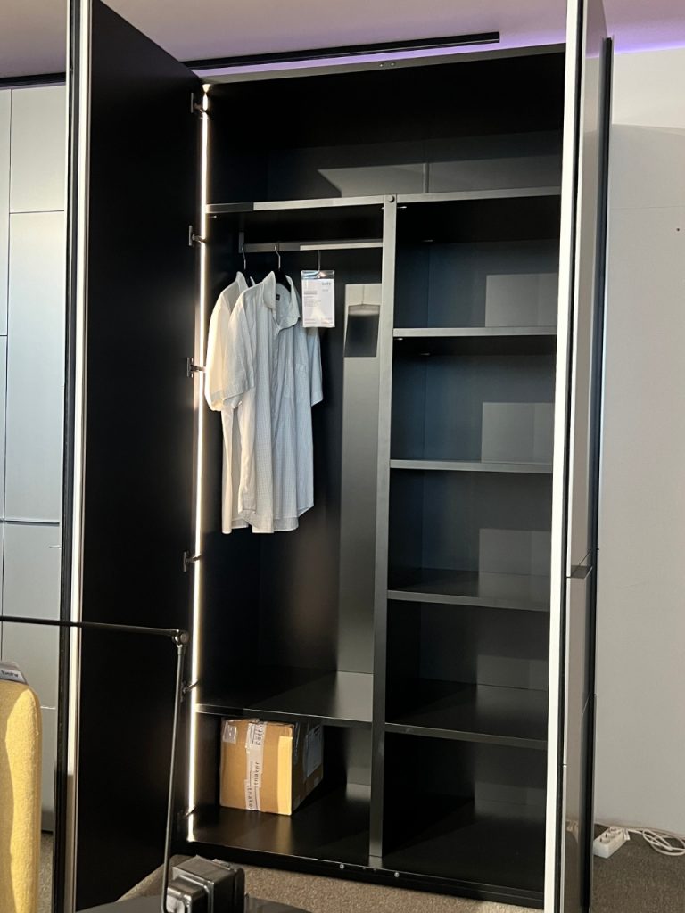 Open black wardrobe with white shirts on a hanging rail, empty shelves on the right, and a small cardboard box on the bottom shelf.