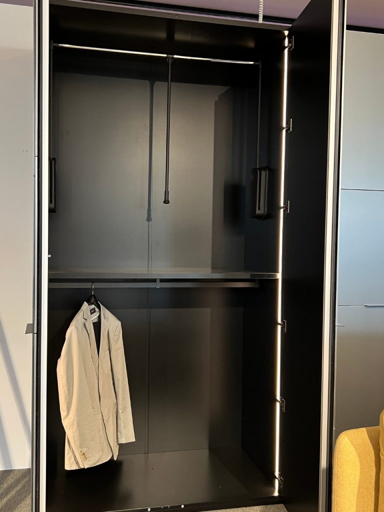 Open dark wardrobe with a beige blazer on a hanger beneath a metal rod; vertical LED lighting runs along the right edge.