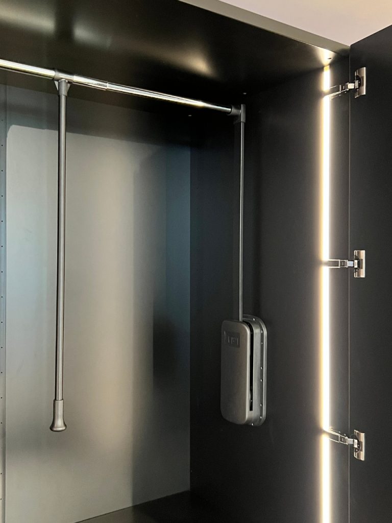 Dark cabinet interior with a metal hanging rod and a vertical LED light strip along the door; a wall-mounted control unit is visible on the back panel