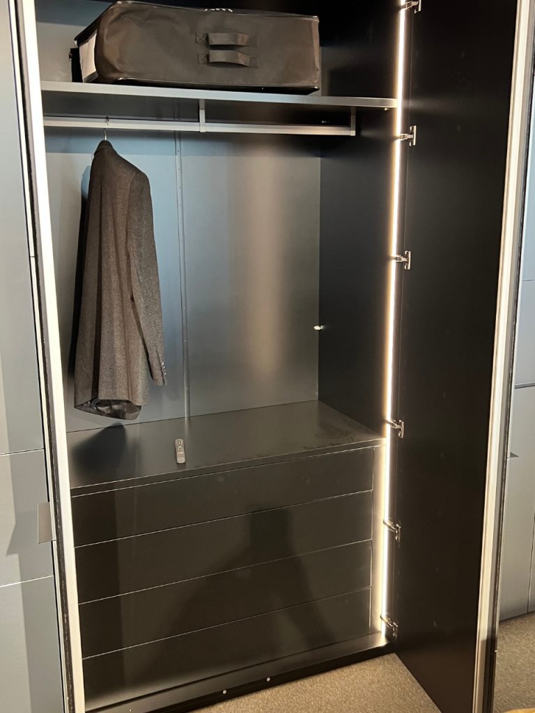 Open black wardrobe with a gray coat on a hanger, a top shelf with a black bag, and four drawers at the bottom; LED edge lighting along the door.