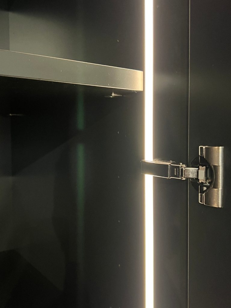 Dark cabinet interior with a vertical LED strip on the right and a metal hinge/lock securing the door; a horizontal latch extends from the hinge mid-door edge for security.