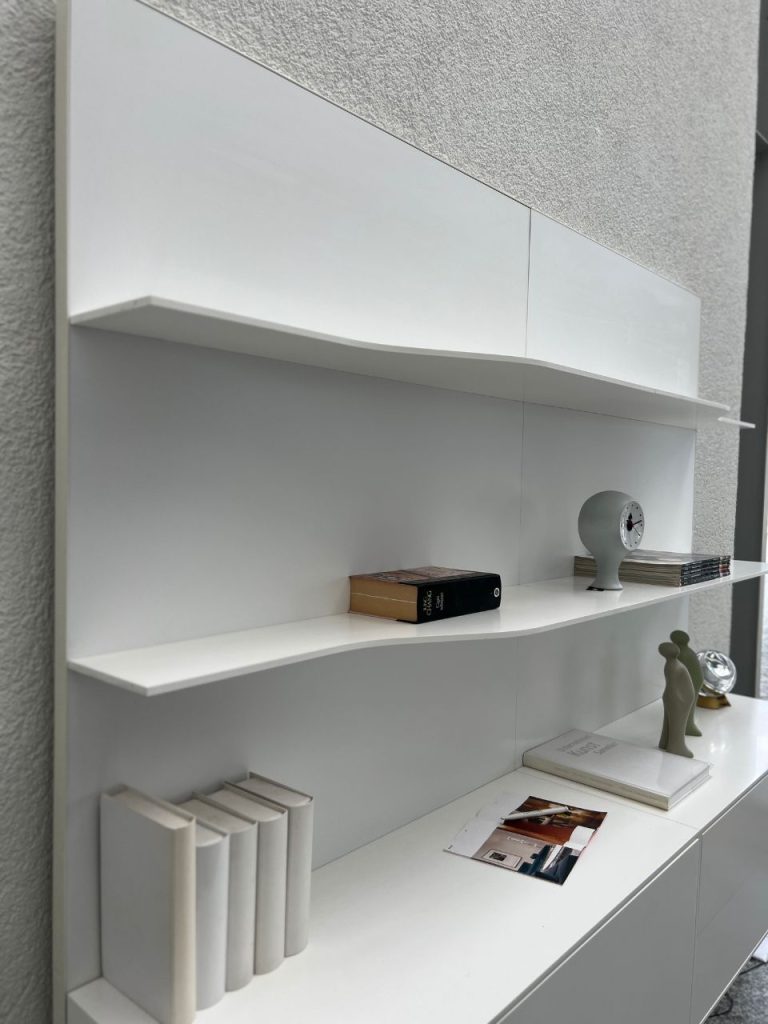 White modern shelving unit with curved edge shelves displaying a few books, a round clock sculpture, magazines, and minimalist figurines on a light wall.
