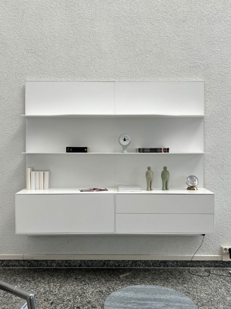 Minimalist white storage wall unit with open shelves and two lower drawers, mounted on a textured light wall; decor includes a small clock, books, two abstract figurines, a glass globe, and magazines on the shelves.