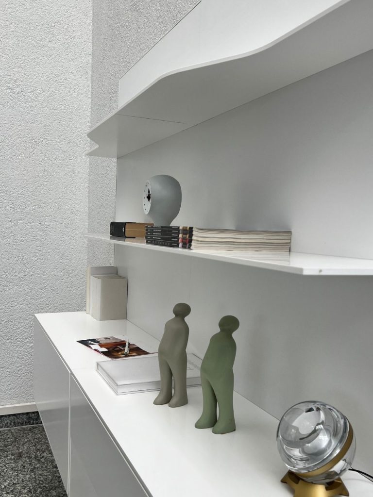 White modern shelf with decorative gray figurines, magazines, and a glass orb lamp on a glossy cabinet.