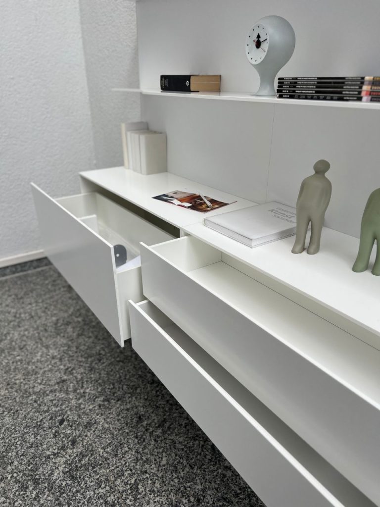 White modular cabinet with open drawers on a speckled carpet; on top are a clock sculpture, stacked magazines, and small figurines, with a white book and a magazine on the desktop.