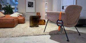 Showroom living room: brown leather sofa, pink chair with orange BEHR sale tag, shag rug, and glass tables.