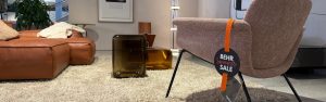 Showroom living room with a brown leather sectional, cream rug, and a beige fabric chair with an orange BEHR Summer Sale tag hanging from the arm.
