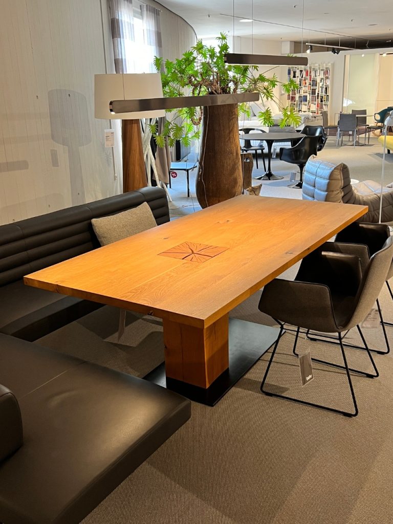 Bright corporate lounge with a large wooden table, black leather bench seating, and a tall potted tree under suspended lights.
