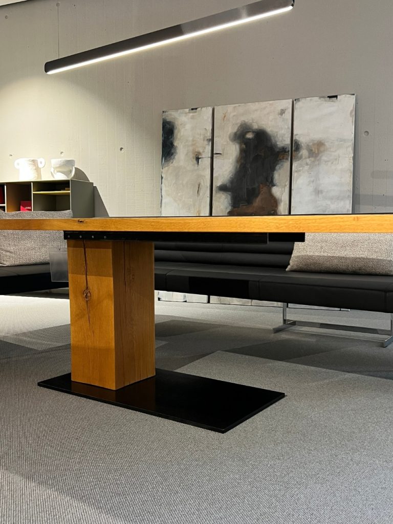 Modern showroom scene with a wooden-top table on a thick rectangular pedestal and a black base, backed by a gray sectional and abstract triptych on the wall.