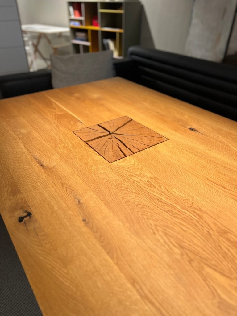 Wooden table with a square wood plug in the center showing cracks, background shelves and seating are out of focus (office/lounge setting).