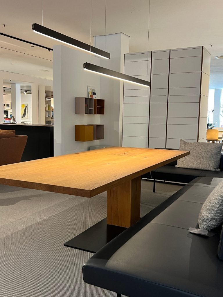 Modern showroom lounge with a long wooden table, gray sectional seating, and suspended linear lights above in a bright, open space.