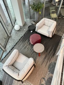 Top-down view of a modern lounge with two striped armchairs, a red pouf, small round tables, and a potted plant by glass walls.