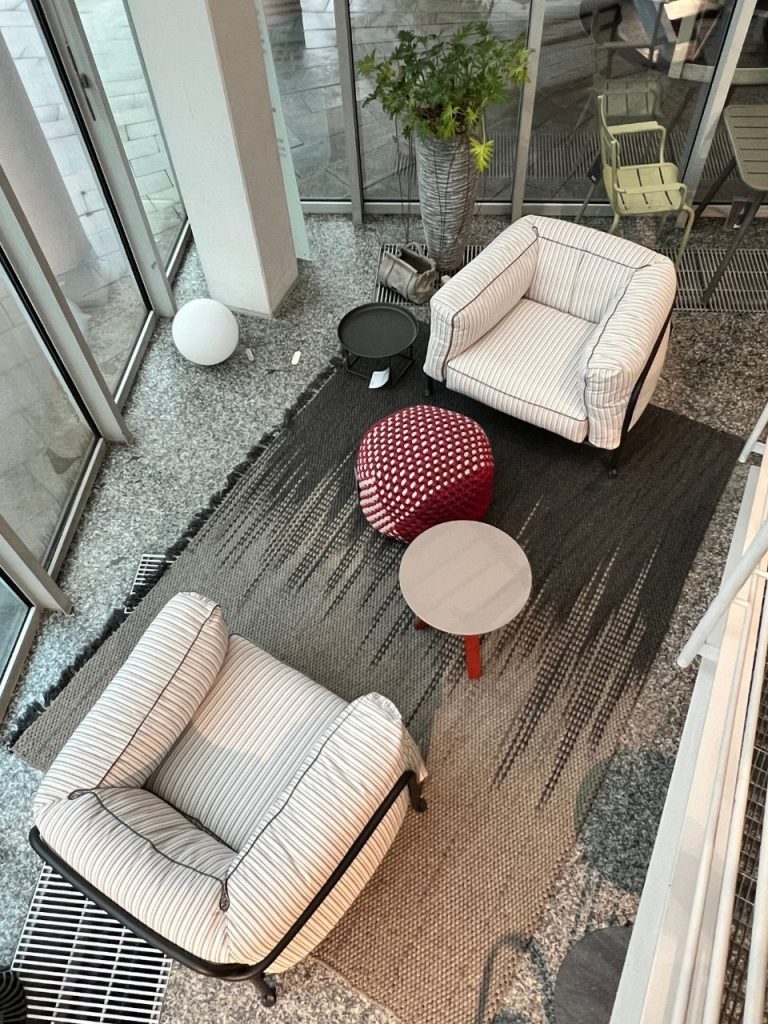 Top-down view of a modern lounge with two striped armchairs, a red pouf, small round tables, and a potted plant by glass walls.