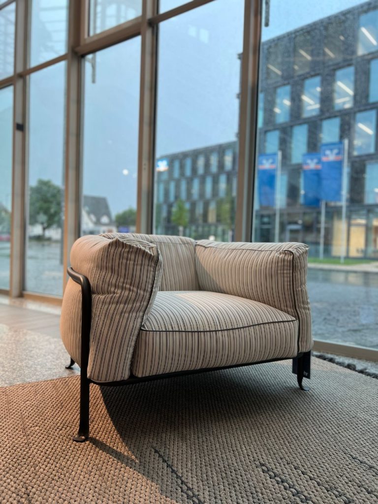 Striped upholstered armchair with metal legs on a woven rug beside tall windows, overlooking a rainy urban street outside the showroom.