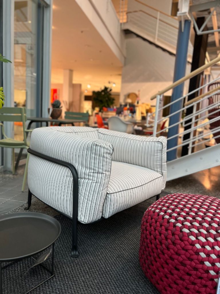 White and gray striped armchair with black metal frame in a modern indoor lounge, next to a red knit pouf and a small round table on a dark rug.