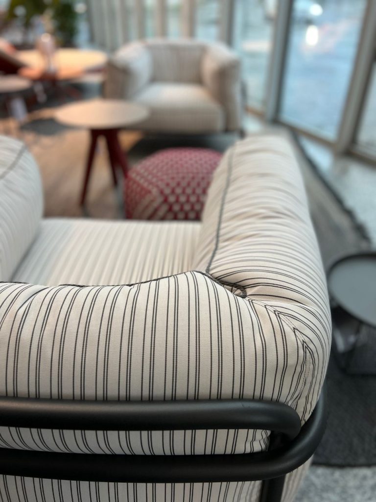 Close-up of a cream-colored striped sofa armrest in a bright lounge with round tables and additional seating in the background through glass walls.
