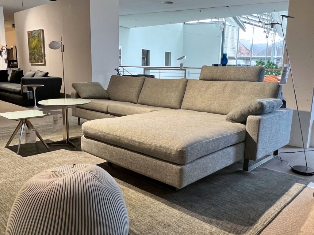 Large light-gray sectional sofa with chaise in a bright showroom, with a round white coffee table nearby.