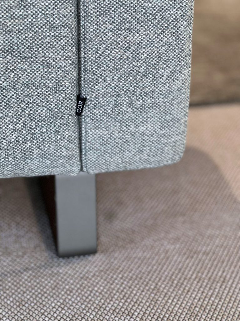 Close-up of grey textured upholstery on a sofa corner with a small black tag reading 'COR' and a metal leg nearby
