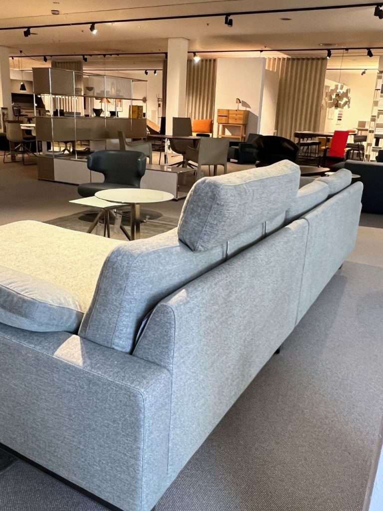 Open-plan showroom with a light gray sectional sofa, a white round coffee table, and various chairs in the background.