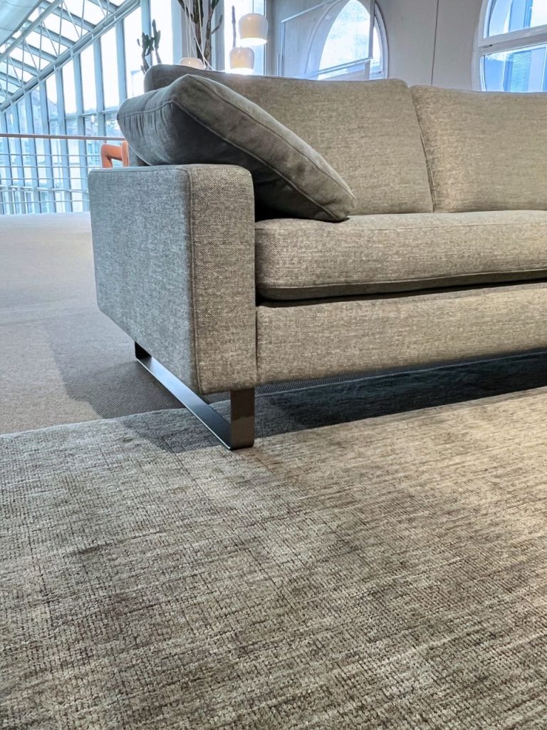 Grey fabric sofa with a tilted back cushion in a modern lobby, metal legs, and large windows in the background.