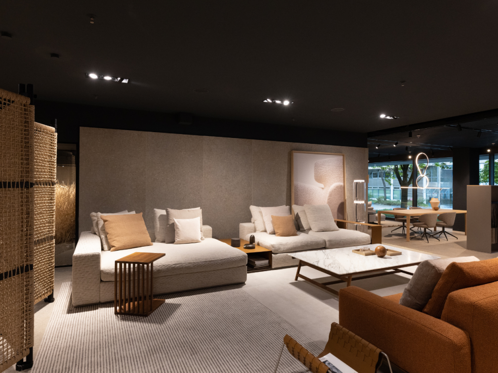 Modern showroom living area with beige sectional sofas, neutral pillows, and a marble coffee table.
