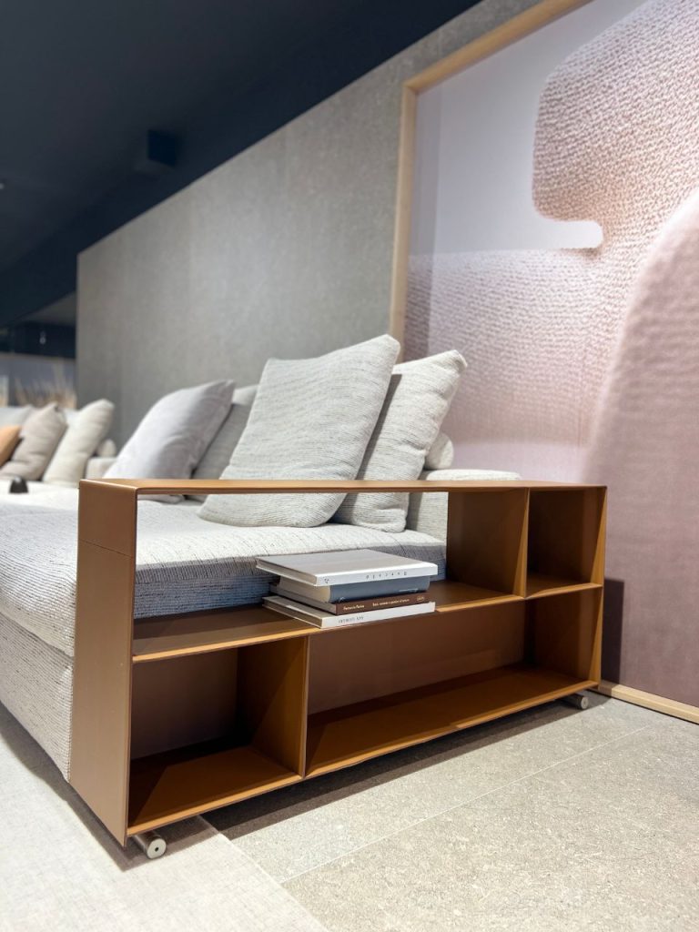 Brown modular sofa-side console with open cubbies and a stack of books beside a gray upholstered sofa in a showroom.