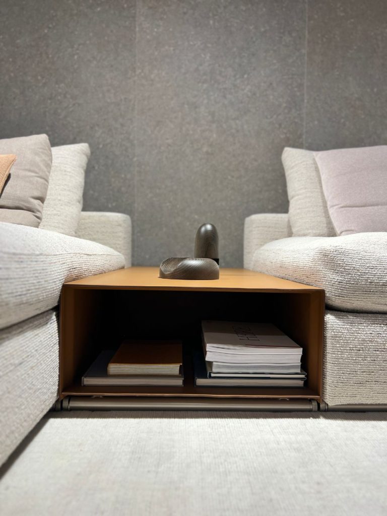 Low brown side table between two gray armchairs, with a dark curved sculpture on top and books stored in the lower shelves.
