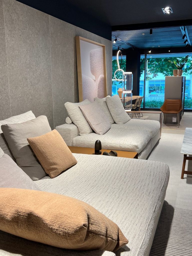 Modern showroom scene with a light gray sectional sofa, beige pillows, and a camel-colored throw across a contemporary living room setting.