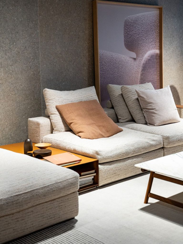 Beige fabric sectional sofa with multiple cushions and a brown throw pillow, beside an orange side table holding magazines and small decor pieces, a framed purple artwork against a gray wall behind.