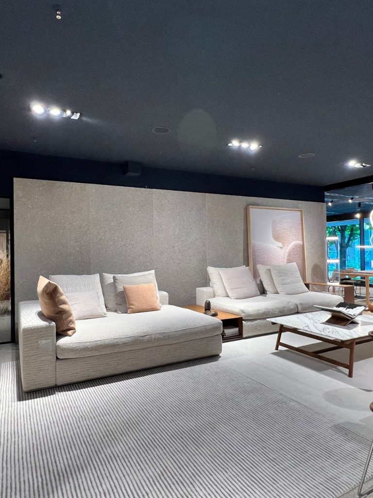 Modern showroom living room with two beige sofas, neutral cushions, a marble coffee table, and a striped rug.