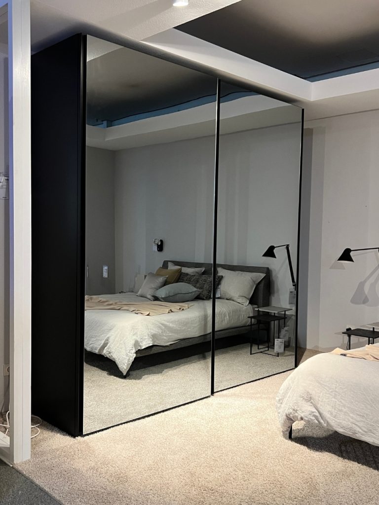 Bedroom with a large black-framed mirrored wardrobe reflecting a white-blanket bed and matching nightstands with black task lamps on gray walls and beige carpet.